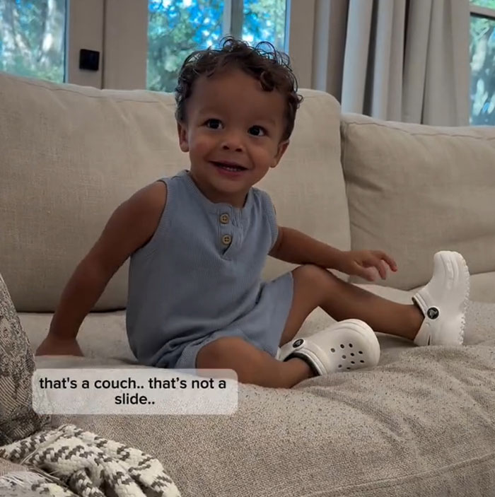 Toddler from 'Okay Baby' smiling on a couch, wearing a blue outfit and white shoes.