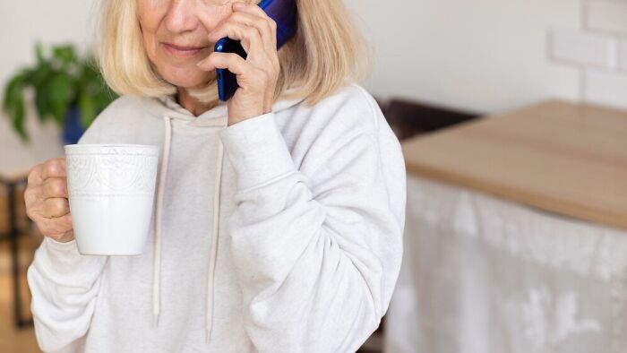 Woman talking on the phone, holding a mug, casually dressed in a light hoodie at home.
