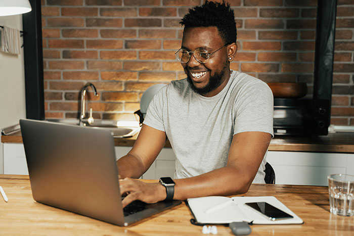 Man smiling at laptop in kitchen, planning an expensive trip to visit long-distance girlfriend.