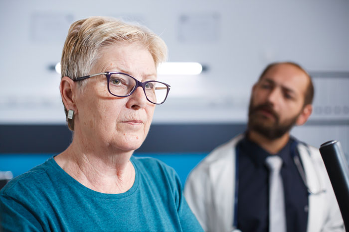 Elderly patient ignored by inattentive doctor, with concerned nurse and upset wife in the background.