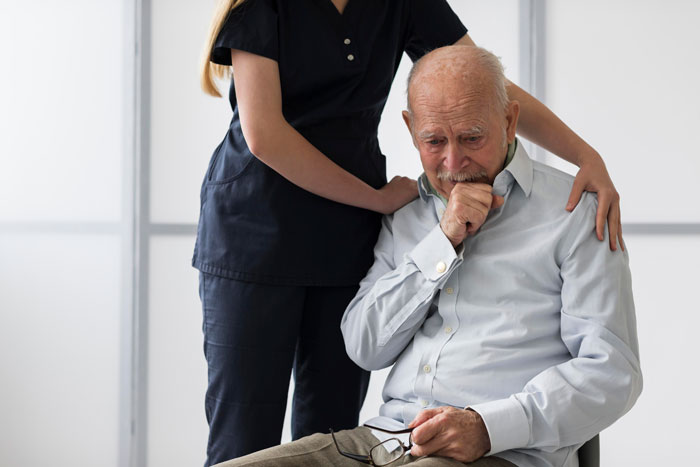Elderly patient sitting, comforted by a nurse, looking contemplative in a clinical setting.