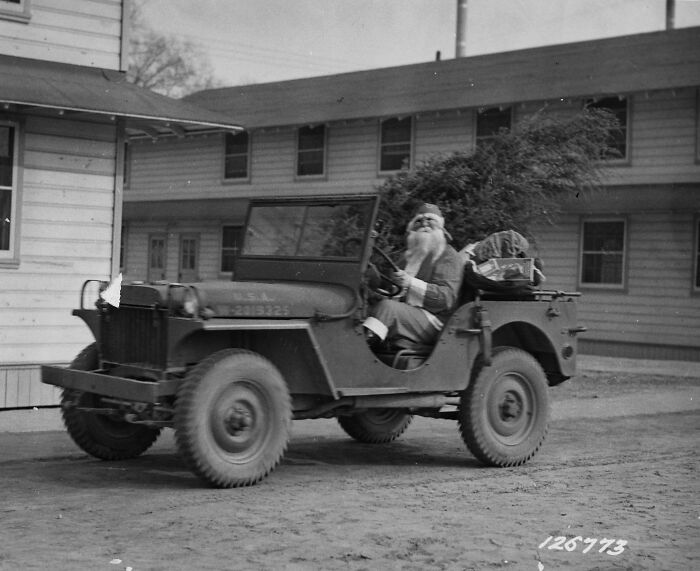 Santa driving a military jeep during WWII, with parcels in the back, in front of a barrack-style building.