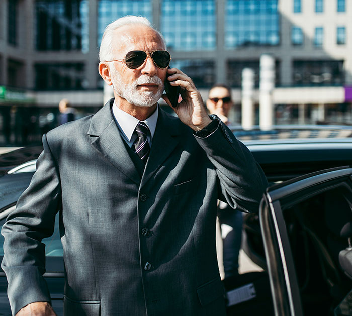 Elderly man in a suit with sunglasses, on the phone, exiting a luxury car, representing out-of-touch wealthy lifestyle.