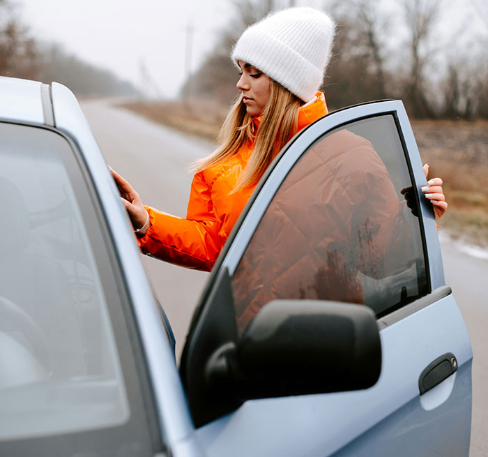 Woman in bright orange jacket and white beanie, opening a car door on a rural road.