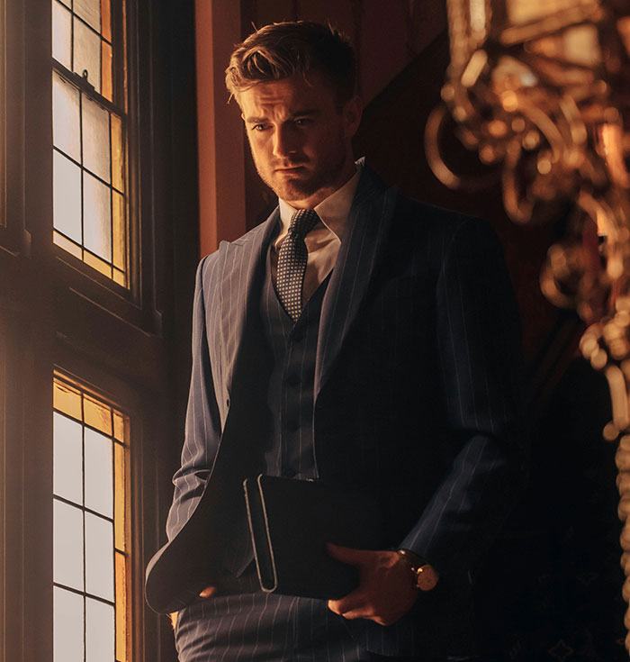 Man in a striped suit by a window, conveying out-of-touch wealth through a contemplative pose.