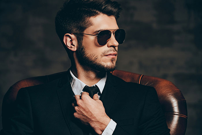 Man in a suit and sunglasses adjusting tie, sitting confidently, symbolizing out-of-touch wealthy lifestyle.