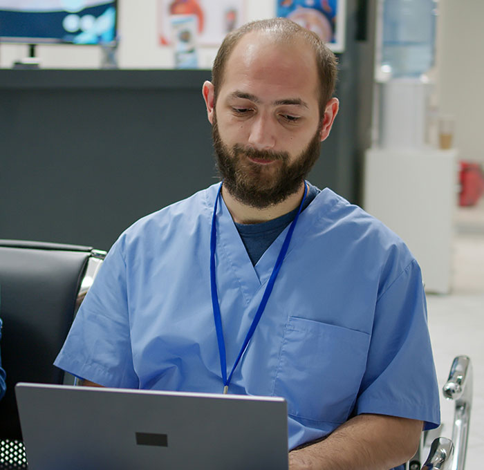 Bearded man in blue scrubs sitting with a laptop, likely pondering out-of-touch statements by the wealthy.