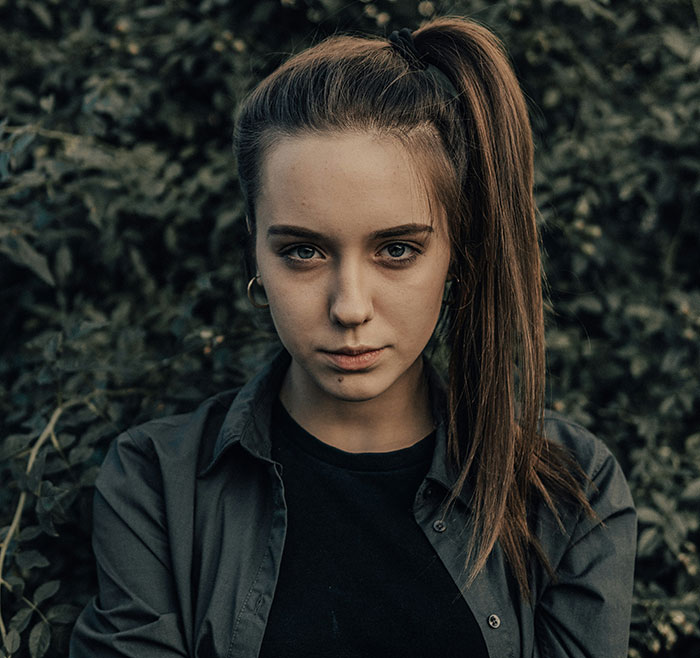 Young woman with a ponytail and hoop earrings, wearing a dark shirt, against a leafy background, representing a candid moment.