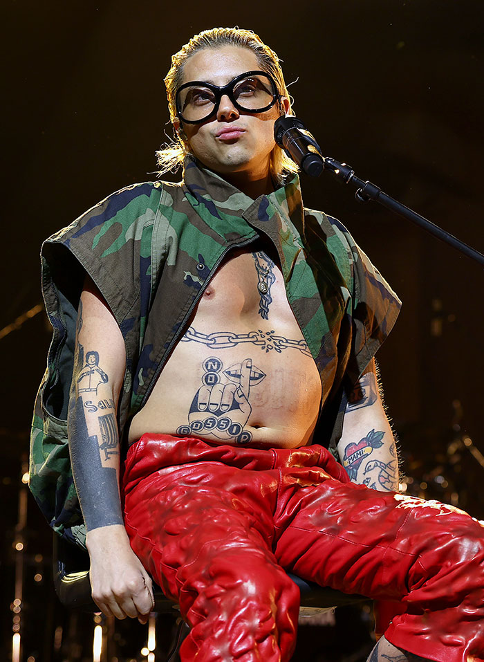 Celebrity in bold outfit at Coachella 2025, wearing oversized glasses, camo vest, and red pants, making a statement on stage.
