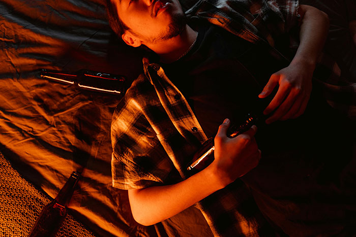 Person lying on a blanket, holding a bottle, surrounded by beer bottles, in a dimly lit setting related to experiences at 18.