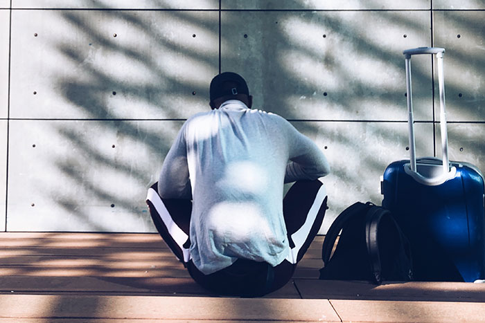 Person sitting against a wall with suitcase and backpack, reflecting on parents kicking kids out at 18.
