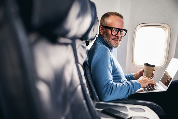 Man in airplane seat with coffee and laptop, smiling, embodying a relaxed passenger experience.