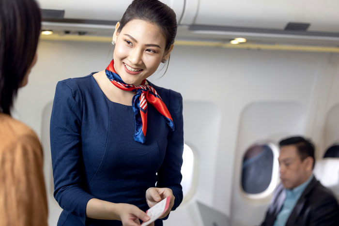 Flight attendant smiling while handling ticket, passenger reading nearby; highlighting travel experience.