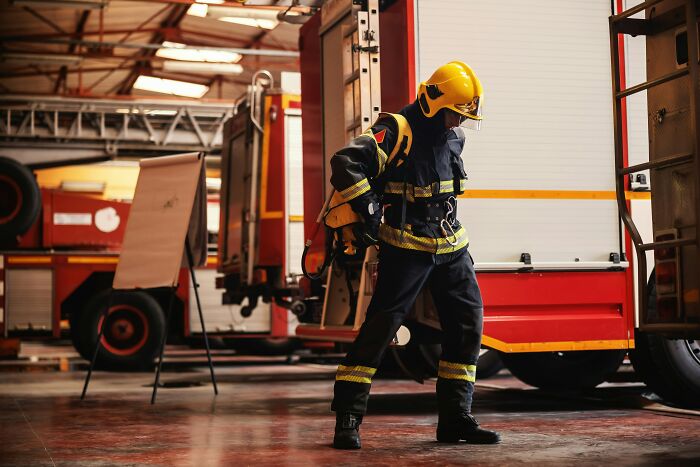 A firefighter in uniform stands in a garage with fire trucks, adjusting gear.