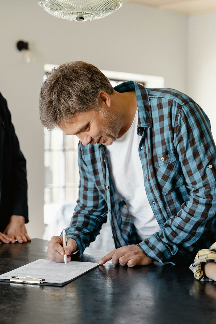 Man signing a document, possibly about pets, wearing a plaid shirt in a well-lit room.