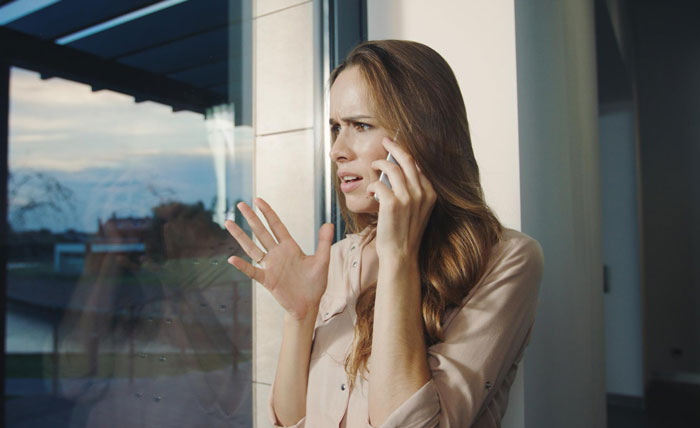 Woman looking concerned on a phone call, standing by a window, discussing strangers claiming to be half-siblings.
