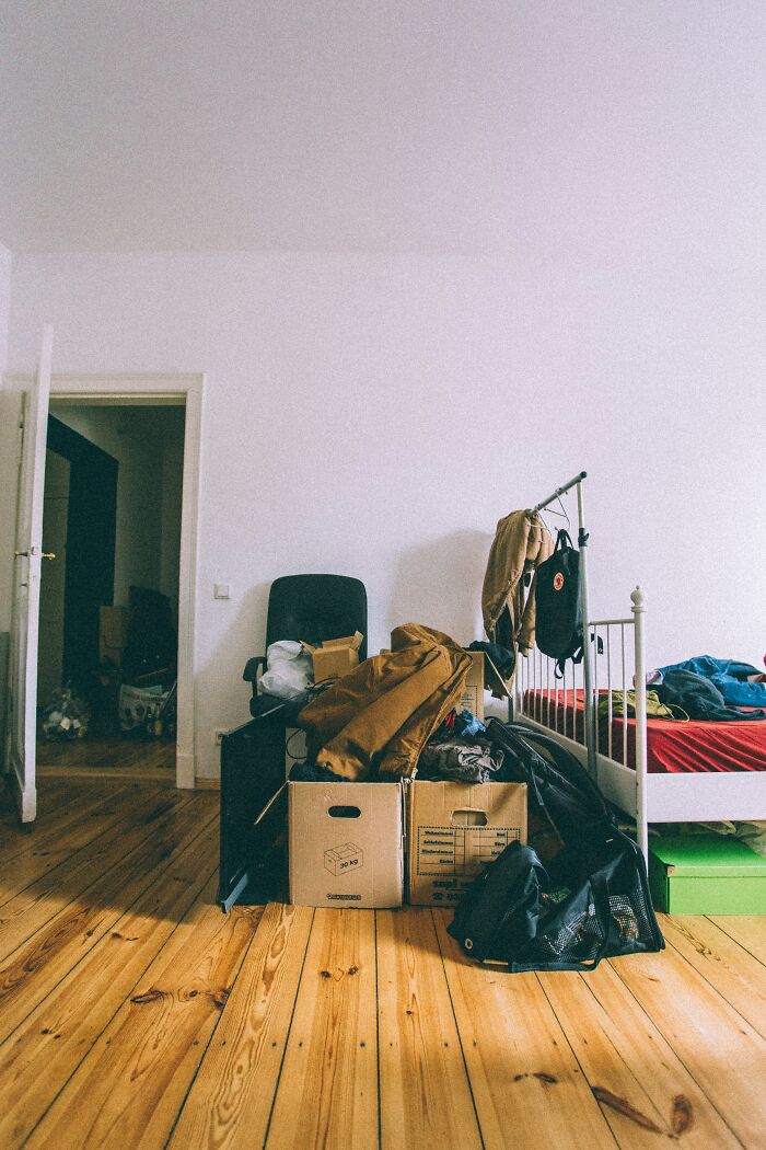 Cluttered room resembling a glitch in the matrix, with boxes and clothes piled beside a bed.