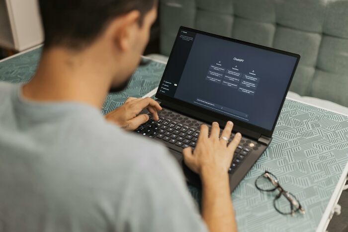 Person using ChatGPT on a laptop in a casual setting, with glasses placed on the table nearby.