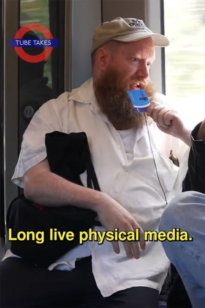 A person on a train discussing physical media, captioned &ldquo;Long live physical media,&rdquo; wearing a cap and holding a microphone.