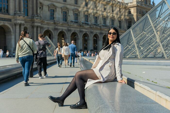 A woman in sunglasses and a white coat sitting outside a grand building, portraying a luxurious lifestyle.