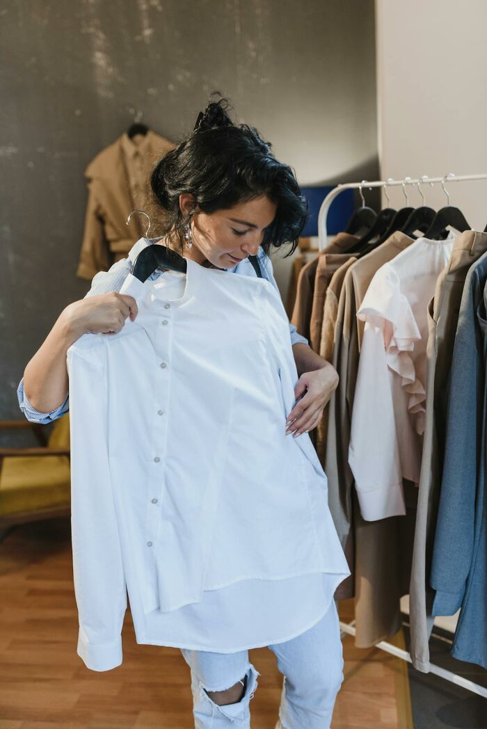Woman holding and examining a white shirt in a clothing store with various brown and white garments on racks behind her.