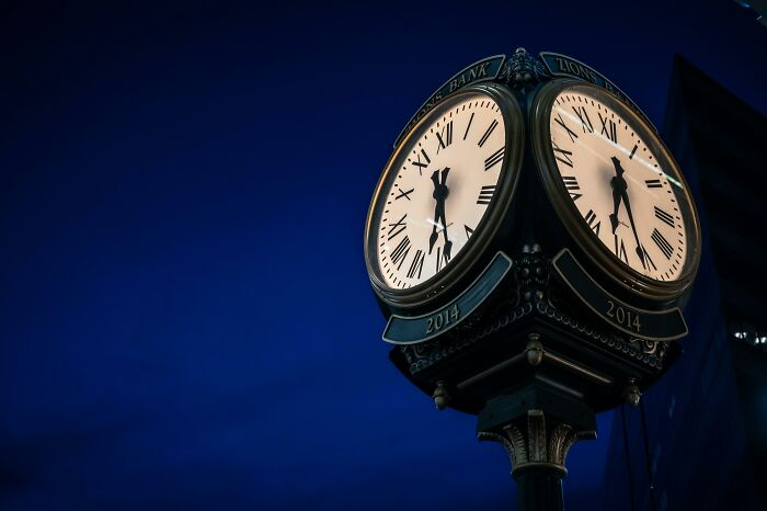 Vintage street clock showing Roman numerals glowing against dark blue evening sky on a black post.
