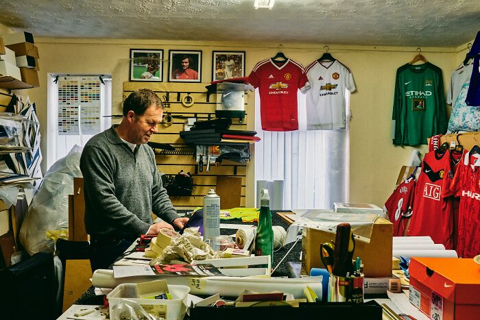 Man in a cluttered office with soccer jerseys and framed photos on the wall, focusing on paperwork.