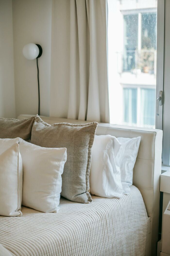 Neatly arranged pillows on a bed by a window with light curtains, creating a cozy atmosphere.