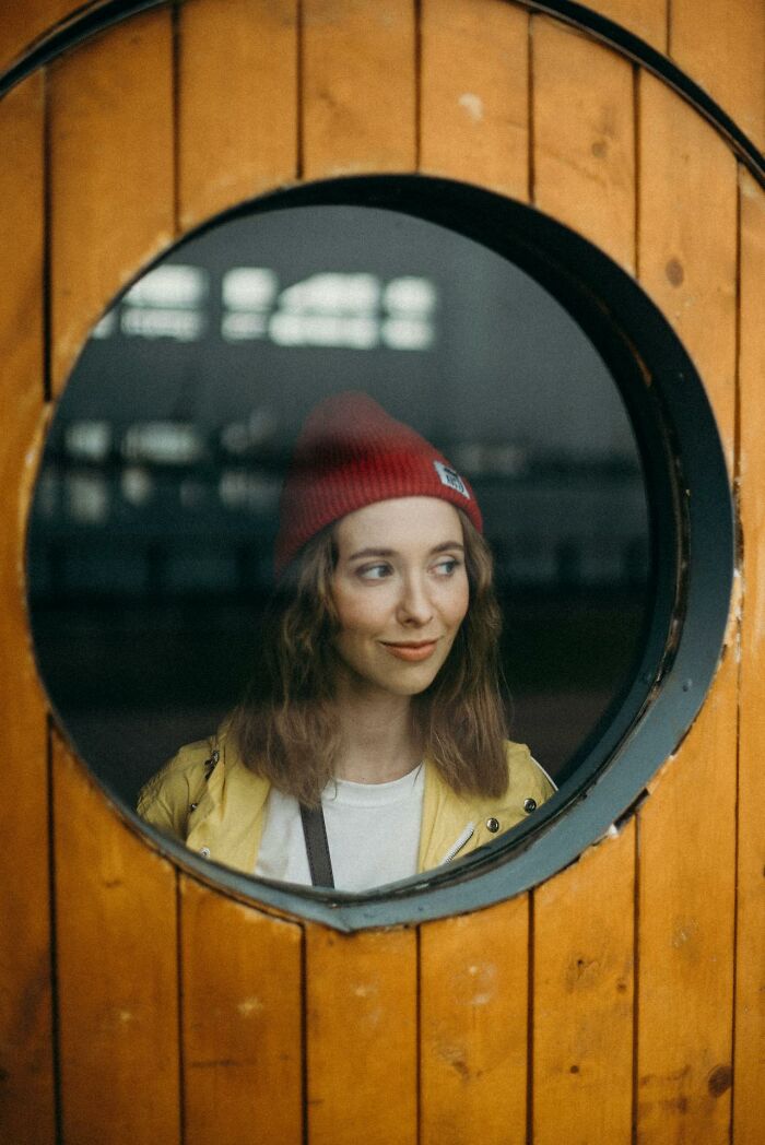 Woman in red beanie behind a round window, evoking a vessel-like aesthetic.