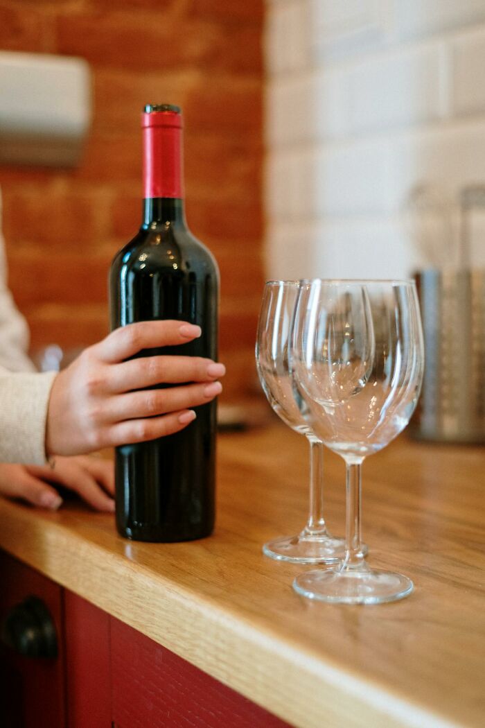 A person holding a wine bottle next to empty glasses on a wooden counter indoors.