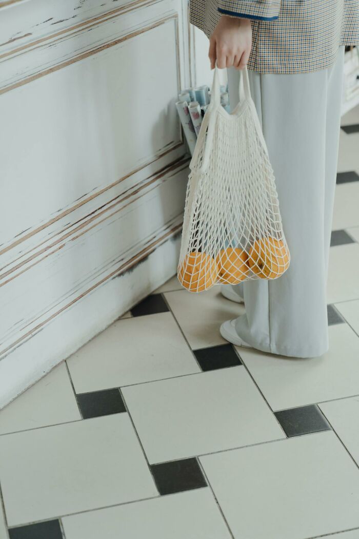 Person holding mesh bag with oranges on checkered floor, beside stylish cabinet.