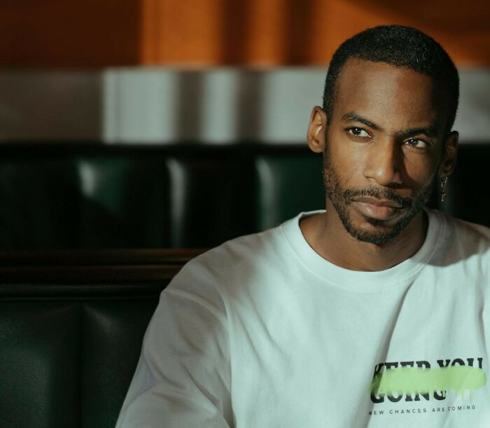 Man seated in dimly lit room, wearing a white shirt, contemplating.