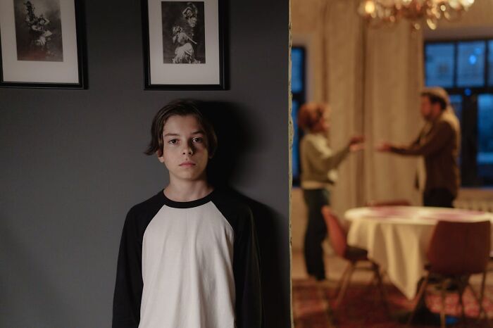 A young boy stands solemnly in the foreground, while two adults argue in the blurred background, highlighting trauma.