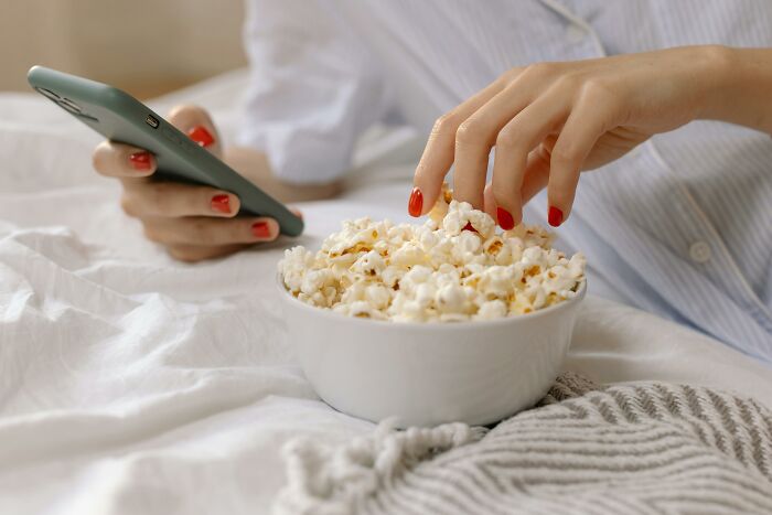 Person with red nails holds a phone and eats popcorn from a bowl, searching for a movie.