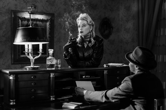 Black and white scene of a classic film noir setting with a woman smoking and a man in a fedora at a desk. Black and white scene of a classic film noir setting with a woman smoking and a man in a fedora at a desk.