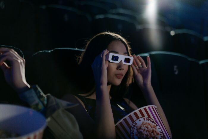 Young woman wearing 3D glasses and holding popcorn while watching a movie in a dark theater for movie title quiz.