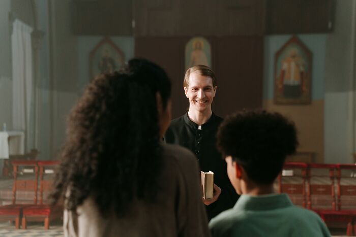 A smiling pastor holding a Bible speaking to church attendees in a dimly lit church setting.