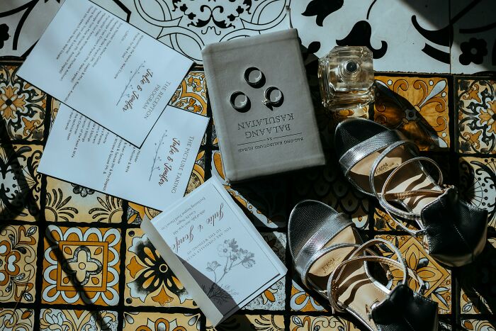 Wedding arrangement with shoes, rings, invitations, and perfume on patterned floor, signifying bride's selections.