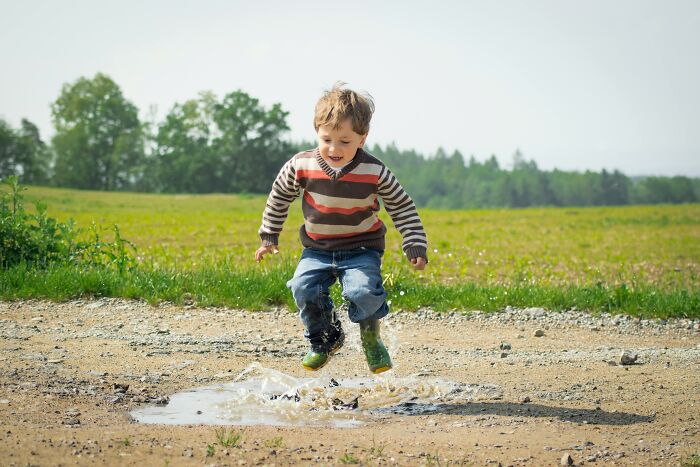 Child joyfully splashing in a puddle outdoors, wearing a striped sweater and jeans, capturing immature playfulness.