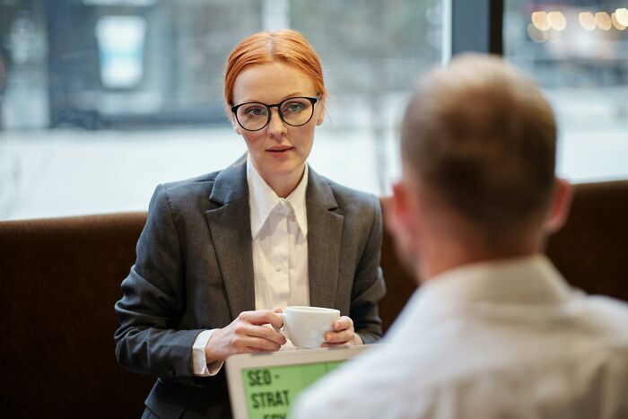 Person in business attire holding a cup, seated across from another person during a job interview discussion.