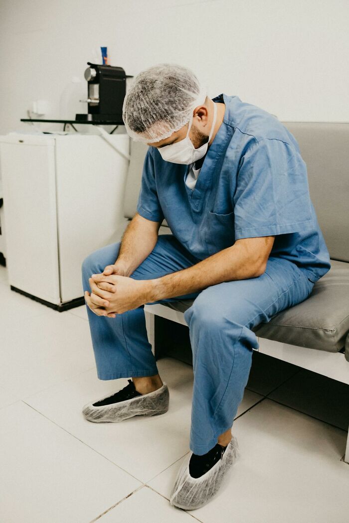 A doctor in scrubs and mask sitting pensively on a bench in a clinical setting.