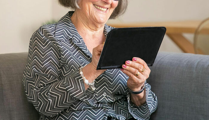 Smiling elderly woman in a patterned blouse using a tablet while sitting on a gray sofa.