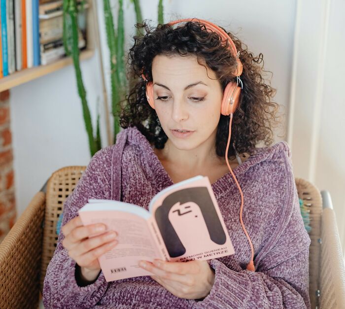 Woman in cozy sweater with headphones reads book, enjoying an affordable treat.
