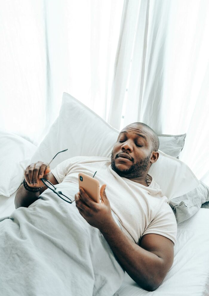 Man in bed holding smartphone and glasses, contemplating something more traumatizing than realized.