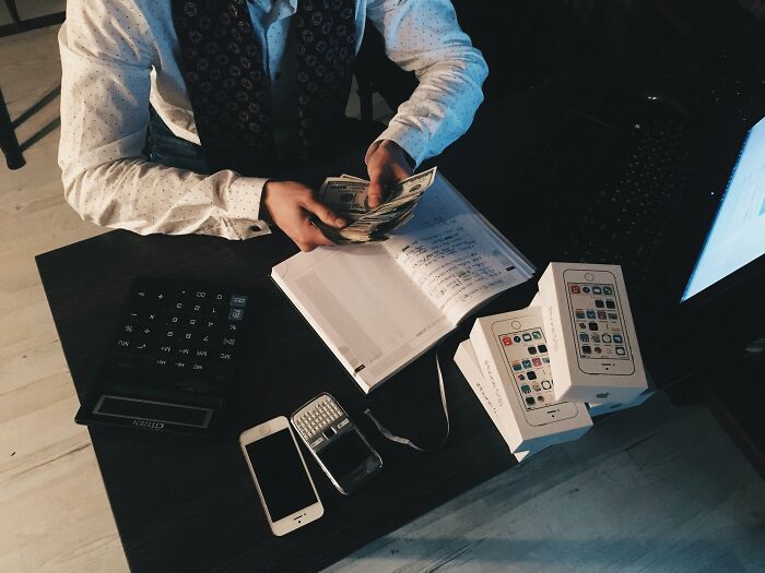 Person handling cash at a desk with calculator, phones, and boxes, illustrating pretending to be rich.