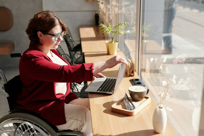 Woman in wheelchair using laptop by window sharing ladies red pill stories with coffee on table in cozy cafe setting