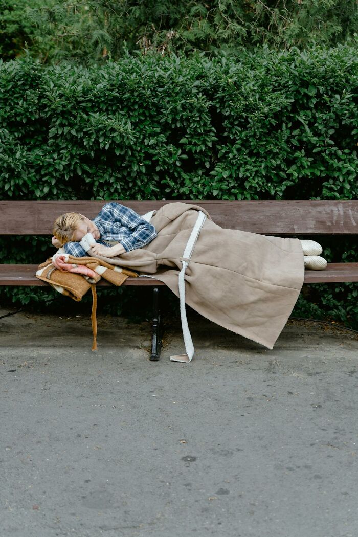 Individual sleeping on a park bench covered with a blanket, creating a somber urban scene.