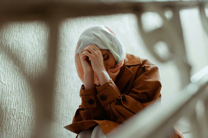 Woman in a headscarf and coat sitting with hands on her head, reflecting common assumptions about women.