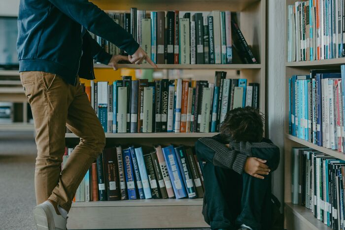 A person points at another sitting against bookshelves, capturing a scene of bullying.