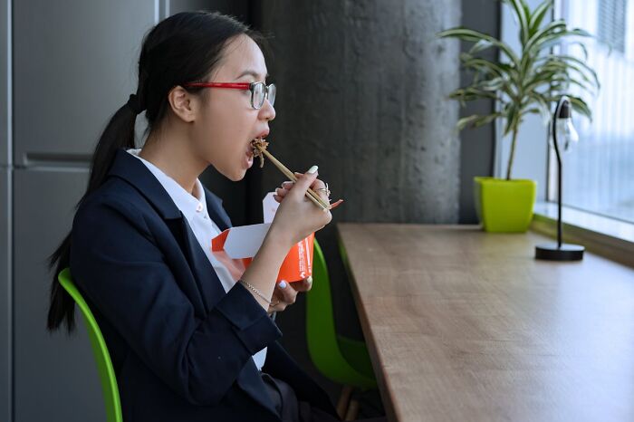 Young woman eating restaurant food with chopsticks from a takeaway box while seated at a wooden table.